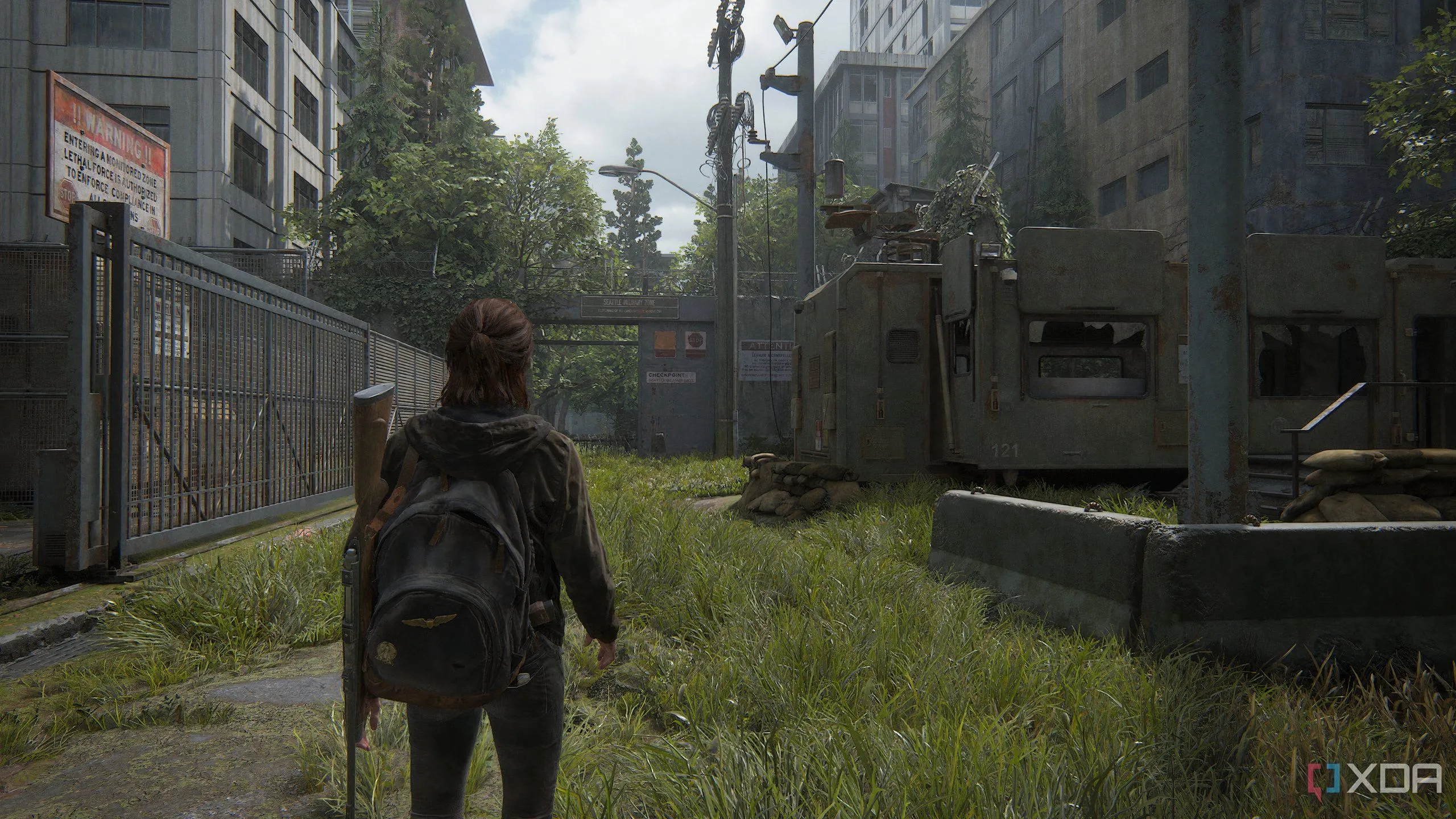 Một cảnh chụp từ game The Last of Us Part II Remastered trên PC, cho thấy Ellie đi bộ qua một khu căn cứ quân sự, làm nổi bật chi tiết môi trường và đồ họa sống động của game.
