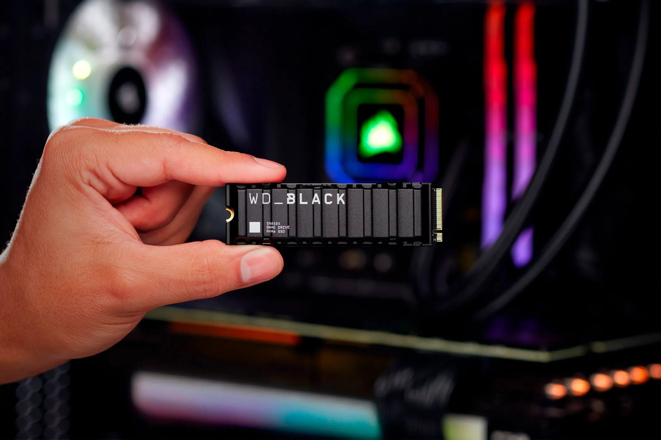 Một người cầm ổ SSD gaming WD Black SN850X với ngón tay cái và ngón trỏ, phía sau là PC gaming có đèn RGB.
