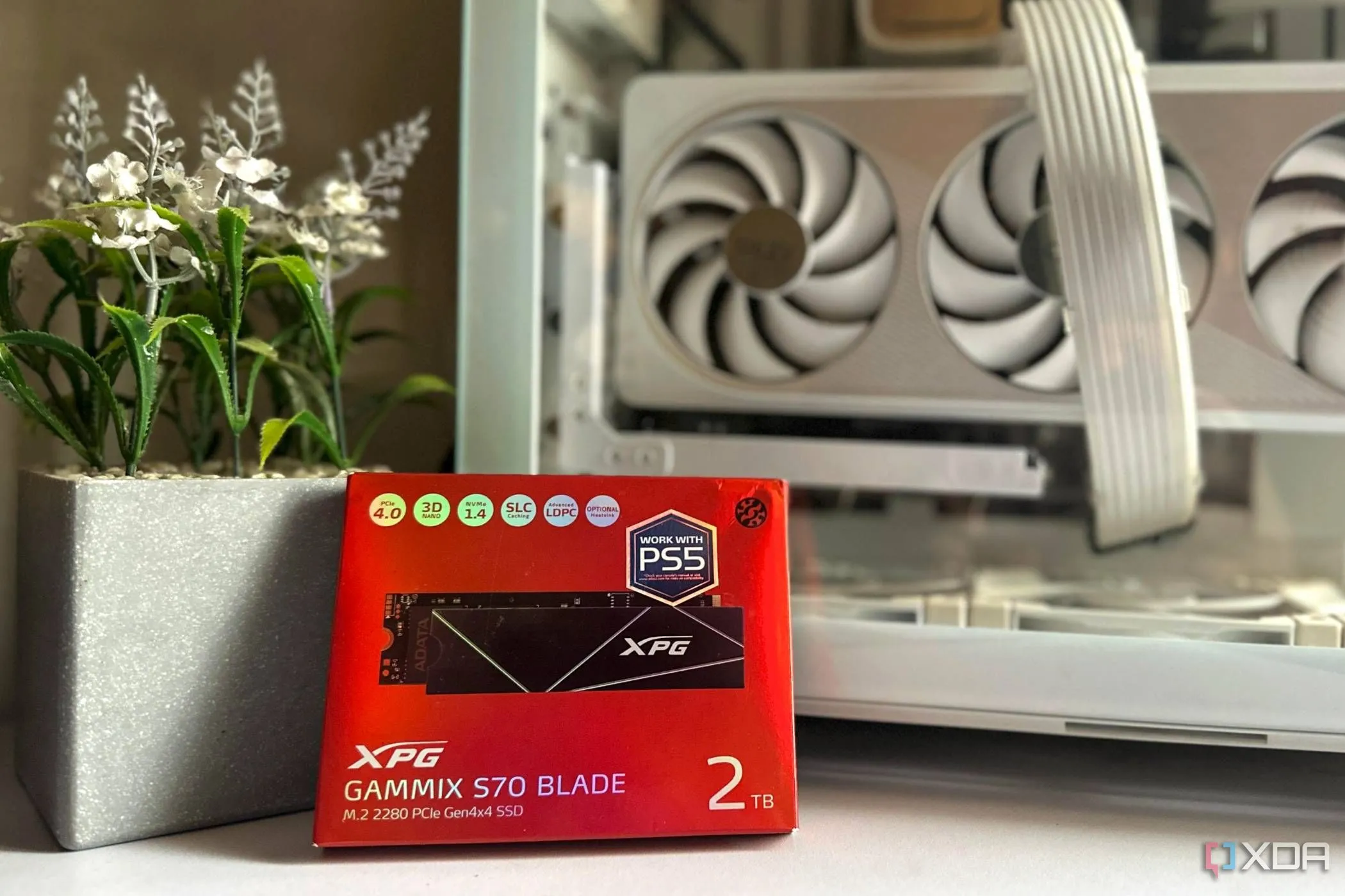 Ổ cứng SSD XPG Gammix S70 Blade 2TB trên hộp sản phẩm, gần chậu cây giả và vỏ case máy tính
